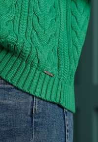 Grüner Kabelstrickpullover mit geripptem Saum und kleinem metallischen Logoakzent. Kombiniert mit blauen Jeans.
