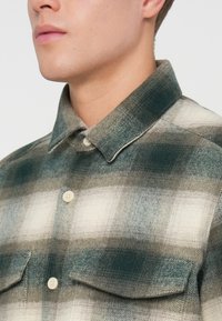 Camisa a cuadros en colores verde, gris y crema. Hecha de franela, con cuello abotonado y dos bolsillos frontales. Tejido texturizado.