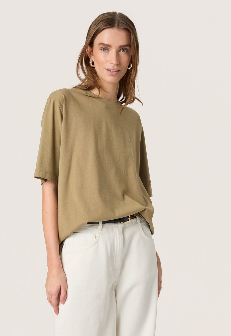 Tricou oversized de culoare verde măslin din bumbac moale, cu guler rotund și mâneci scurte, asociat cu pantaloni albi evazați și o curea neagră.