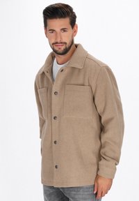 Man met kort donker haar en een baard, gekleed in een beige knoopjas en grijze spijkerbroek, staande tegen een effen witte achtergrond.