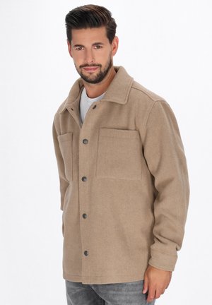 Man with short dark hair and beard wearing beige buttoned coat and gray jeans, standing against plain white background.