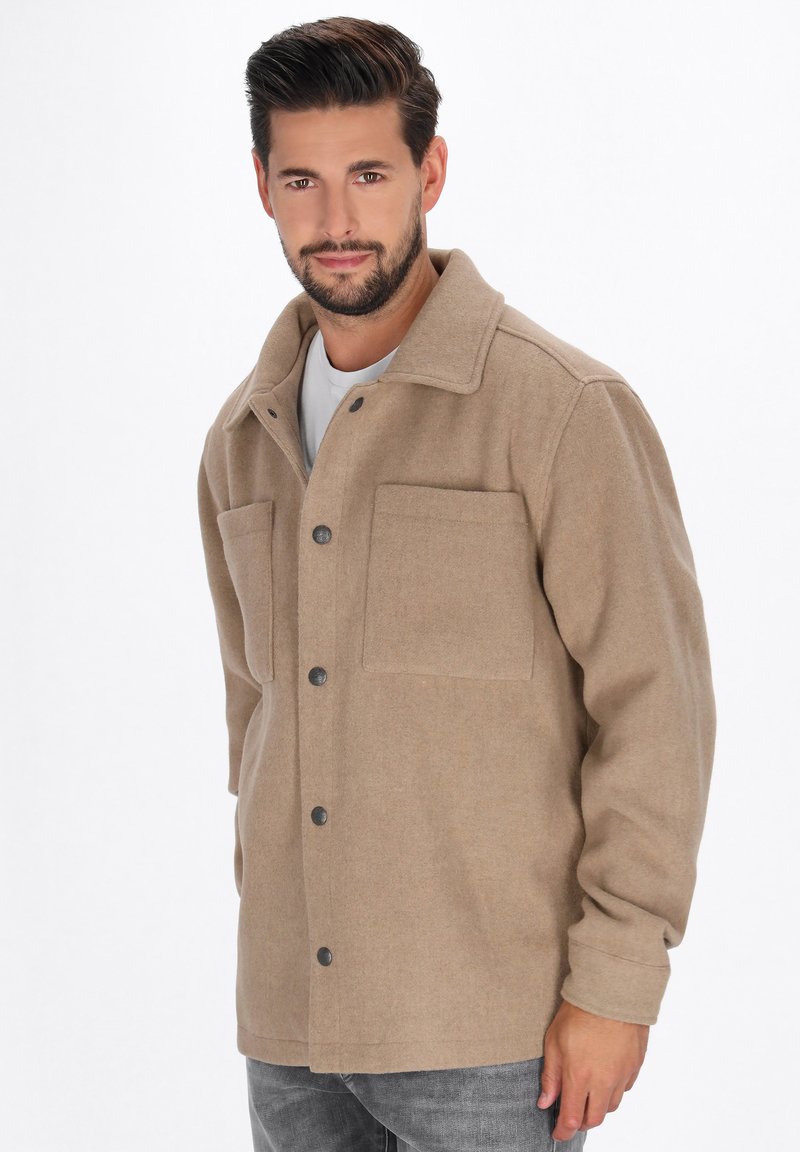 Man met kort donker haar en een baard, gekleed in een beige knoopjas en grijze spijkerbroek, staande tegen een effen witte achtergrond.