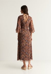 Bruine maxi jurk met een gedrukte print, wijde mouwen, een V-vormige rug en strikdetail. Bevat een geometrisch patroon in aardetinten en een zachter, vloeiend materiaal.