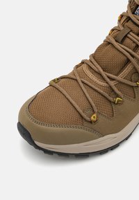 Ruskeat vaelluskengät, joissa on teksturoitu verkkopinta, vahvistettu varvasosa ja kirkkaan keltaiset sekä punaiset nauhat. Varustettu kumipohjalla, joka tarjoaa pidon.