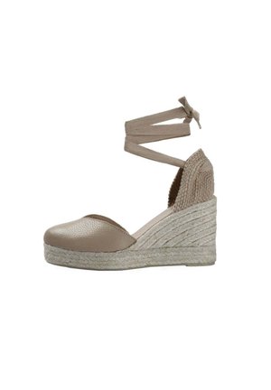Zapato espadrille beige con cuña, puntera cerrada, tacón de cuerda tejida y correa para atar alrededor del tobillo sobre fondo blanco.