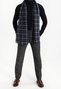 Marinblå rutigt scarf placerat över en svart polotröja, tillsammans med grå texturerade byxor och bruna läderskor, vilket skapar en lager-på-lager look.