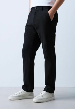 Mann trägt schwarze Slim-Fit-Hose mit Händen in den Taschen und weißen Freizeitsneakern, steht auf hellem Boden vor einfarbigem Hintergrund.