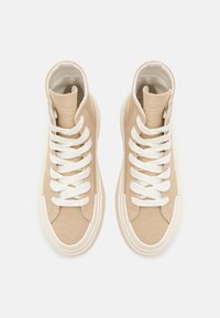 Beżowe trampki typu high-top wykonane z płótna, z białymi sznurowadłami, zaokrąglonym gumowym noskiem oraz kontrastującymi przeszyciami po bokach.