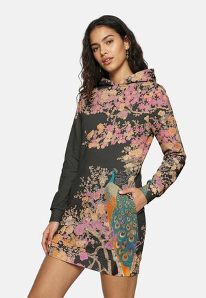 Junge Frau trägt ein schwarzes Kapuzenkleid mit floralen Mustern, verziert mit rosa und orangefarbenen Blüten sowie einem bunten Pfau-Design, die Hand in der Tasche.