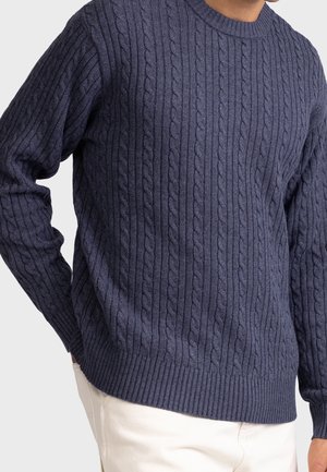 Homme portant un pull torsadé bleu marine avec poignets et ourlet côtelés, associé à un pantalon blanc cassé, mains reposant sur les côtés.
