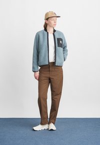 Világoskék, fekete díszítésű boleró dzseki, mellzsebből álló fekete díszítéssel, barna nadrágokkal és fehér tornacipőkkel párosítva. Logóval díszített sapka.