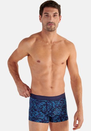 Man with short dark hair wearing blue patterned boxer briefs stands against a plain light background.