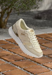 Μπεζ αθλητικά παπούτσια με υφή, διαθέτουν λείο δέρμα, λεπτομέρειες από δίχτυ, λευκή σόλα και μεταλλικά στοιχεία σε χρυσή απόχρωση. Ονομασία μάρκας ορατή.