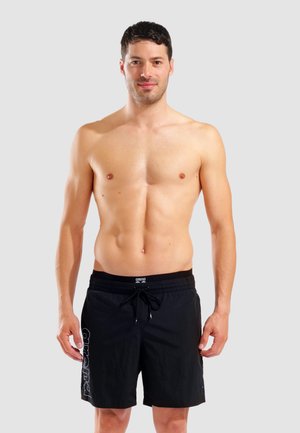 Hemdloser Mann stehend, trägt schwarze Badeshorts mit Kordelzug und "arena" Logo am Bund und am linken Bein, vor einfachem grauem Hintergrund.