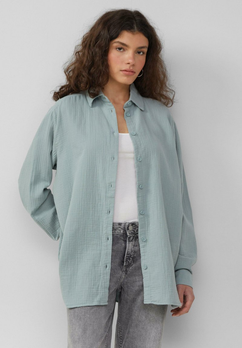 Camisa de botón texturada de color azul claro, con cuello, mangas largas y un corte relajado, llevada sobre una camiseta sin mangas blanca y unos vaqueros grises.