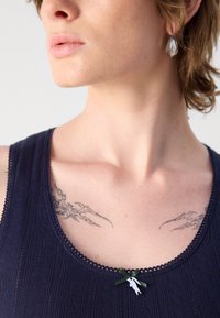 Tamsiai mėlynas tank topas su tekstūruotu audiniu, banguoto kaklo iškirpte ir dekoratyvine žalia juostele su sidabriniu papuošalu krūtinėje. Matomi tatuiruotės.