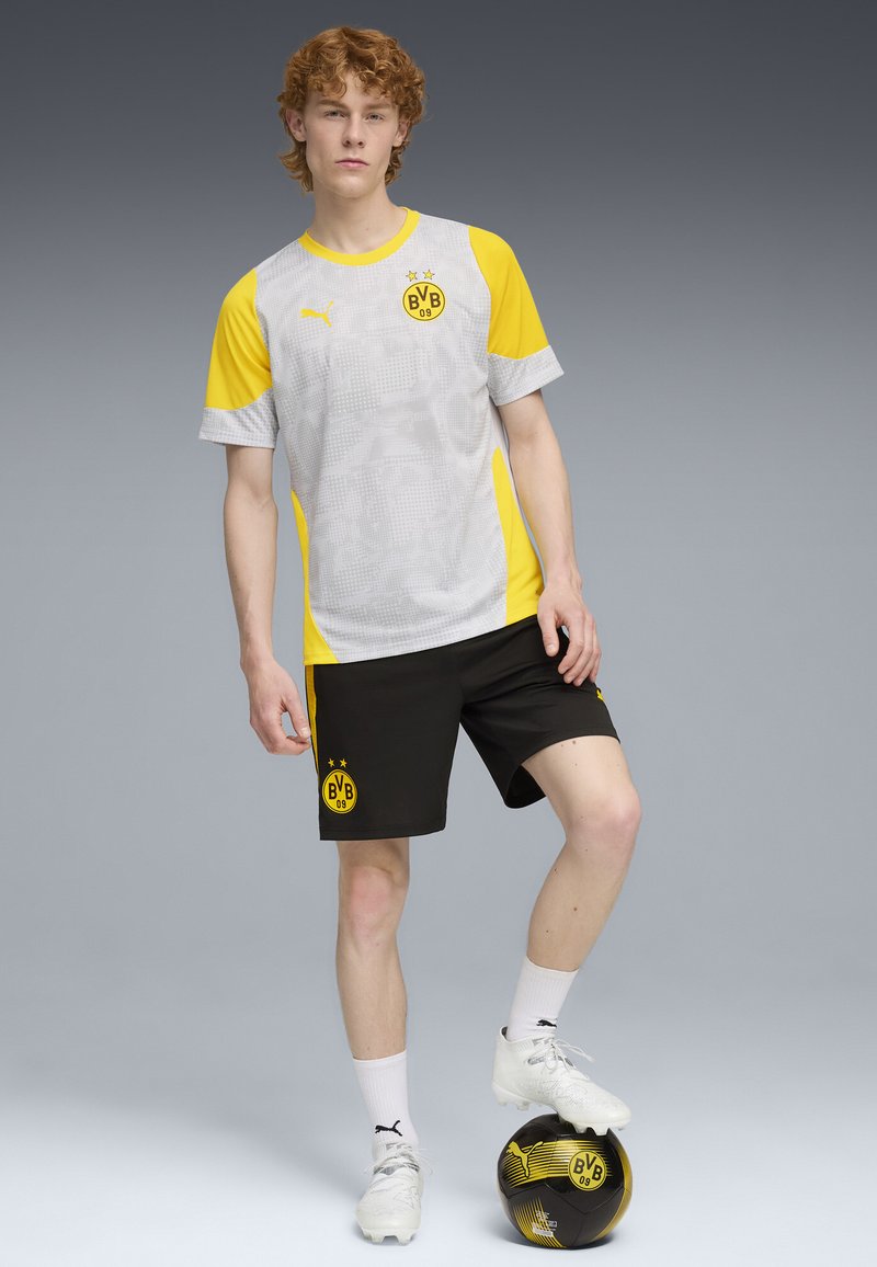 Conjunto deportivo de Borussia Dortmund en negro y amarillo con un patrón gris en la camiseta, mangas cortas, y pantalones cortos negros con logotipos. Un balón de fútbol negro y amarillo descansa a su pie.