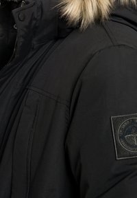 Parka noire avec une capuche bordée de fourrure, dotée d'une coque texturée, d'une fermeture éclair et d'un écusson brodé sur l'épaule gauche.