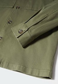 Oliivinvihreä puuvillapaita, jossa on napillinen malli, tasku ja kalvosinnauha yksityiskohtana. Sileä rakenne, klassinen kaulus ja ruskeat napit.