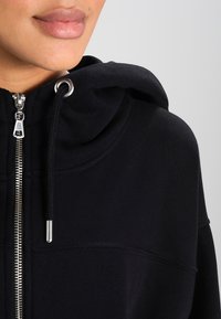Black hoodie with a zip closure and drawstring hood. Features a textured fabric and silver hardware accents. Simple and functional design.