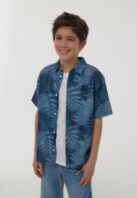 Camicia azzurra a maniche corte con un motivo di foglie tropicali. Indossata sopra una maglietta bianca, abbinata a jeans azzurri chiari.