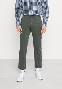 Chinos verde oliva con gamba dritta e una sottile texture, abbinati a una camicia a quadri scura e scarpe da ginnastica bianche.