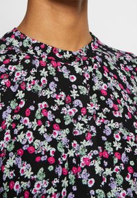 Blouse noire à motif floral avec un col boutonné, ornée de motifs floraux roses, violets et verts sur un tissu texturé.