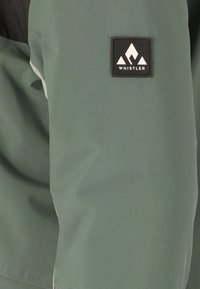 Giacca verde con una texture morbida, caratterizzata da una toppa nera con un logo bianco sulla manica e un accento di colore contrastante.