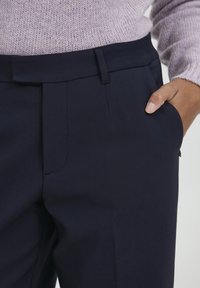 Navy skräddarsydda byxor med en slät yta, enkel veckdetalj och en sidoficka. Midjeband med bältesöglor, stylad med en lätt stickad tröja.
