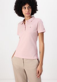 Vaaleanpunainen polo-paita, lyhyet hihat, kaulus, pieni brodeerattu logo rinnassa, pehmeä materiaali, yhdistetty vaaleanbeigeihin housuihin.
