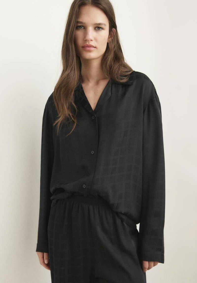 Jongeman met lang bruin haar die een losse zwarte knoopshirt en bijpassende zwarte broek draagt tegen een effen lichte achtergrond.