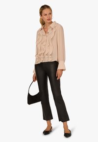 Light beige blouse with ruffled front detail, long sleeves, and button closure, paired with black fitted pants and flat black shoes.