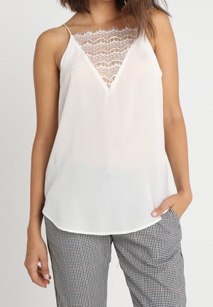 Vrouw draagt een witte mouwloze blouse met kant in de V-hals, gecombineerd met zwart-wit geruite broek, met één hand in de zak.