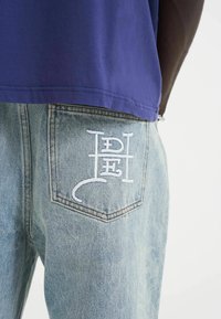 Denim jeans met een lichtblauwe wassings, voorzien van een witte geborduurde logo op de achterzak. Het shirt is paars en heeft een relaxte pasvorm.