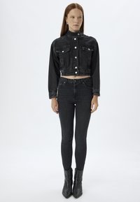 Chaqueta corta de denim negra con bolsillos frontales, cierre de botones y cuello alzado, combinada con jeans negros ajustados y botines.