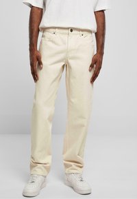 Jean en coton beige avec une coupe droite, dotés de poches avant, d'une fermeture à bouton et d'une surface texturée. Associés à des baskets blanches.