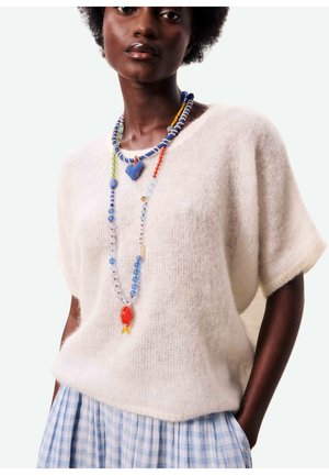 Femme à la peau foncée portant un pull à manches courtes blanc pelucheux, une jupe à carreaux bleue et un long collier coloré en perles avec des pendentifs en forme de cœur et d'ovale.