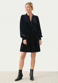Rochie din catifea neagră cu fustă plisată, mâneci lungi și guler cu decupaj. Pur tată cu cizme negre până la gleznă. Textură netedă, până la genunchi.