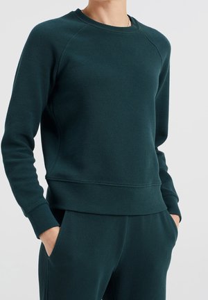 Osoba ubrana w ciemnozieloną, teksturowaną bluzę z długim rękawem oraz pasujące spodnie, z rękami w kieszeniach, widoczna od ramion do bioder.