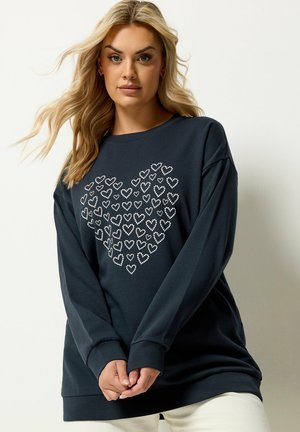 Blonde vrouw in een marineblauwe trui met een hart van kleine witte hartjes, staand tegen een effen lichte achtergrond.