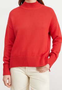 Rode gebreide trui met een hoge geribbelde kraag, lange mouwen en een geribbelde zoom. Gemaakt van zacht stof met een ontspannen pasvorm.