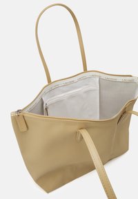 Sac fourre-tout en toile beige avec une fermeture éclair sur le dessus, des anses lisses et une poche intérieure blanche. Présente la marque Lacoste sur la doublure et l'étiquette.