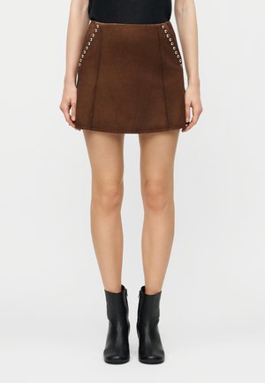 Mini-jupe en daim marron avec détails œillets argentés, portée avec des bottines noires, par une personne debout sur fond blanc.