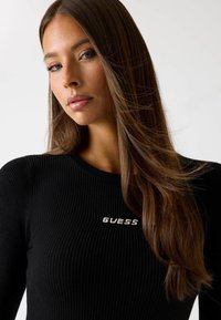 Maglietta nera a maniche lunghe con costine, caratterizzata da un collo rotondo e un logo argento "GUESS" sul petto, realizzata in tessuto liscio e con una vestibilità aderente.