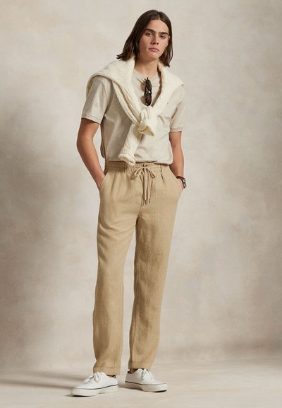 Be�ž lanene hlače z elastičnim pasom, združene s svetlo kremasto majico s kratkimi rokavi in pletenim kremnim puloverjem, ki je zavezano okoli vratu.