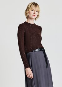 Brauner Pullover mit rundem Halsausschnitt, strukturierten Oberfläche mit bunten Sprenkeln, kombiniert mit hochgeschnittenen grauen Plisseehosen und einem schwarzen Gürtel.