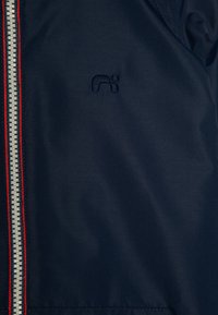 Marinblå vindjacka i strukturerad tyg, röd- och vitrandig dragkedja samt en broderad logotyp på vänster sida.