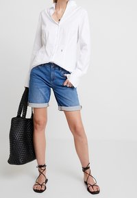 White long-sleeve button-up shirt, blue denim shorts with rolled hem, black woven tote bag, and black strappy sandals.