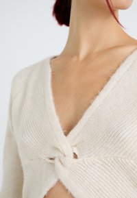 Femme portant un pull en tricot de couleur crème avec un détail de nœud torsadé à l'avant, laissant apparaître une partie de son cou et de sa poitrine.