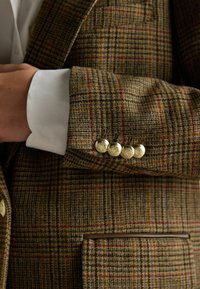 Pruun ja kuldne houndstooth bleiser, millel on kuldsed reljeefsed nupud, tekstureeritud kangas, küljetasku ja nähtav valge särgi mansett.